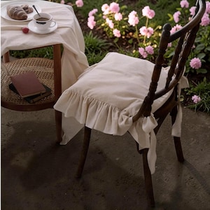 Pode incluir: Cadeira de madeira com almofada creme com borda franzida. Uma pequena mesa está montada com uma xícara de café, um prato com um croissant e um livro. Flores cor-de-rosa ao fundo.