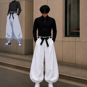 Op de afbeelding: Een zwart shirt met lange mouwen en een kwartritssluiting gecombineerd met een wijde witte broek met een zwarte stoffen riem in de taille. De broek heeft een geplooide zoom bij de enkels en wordt gedragen met witte sneakers.