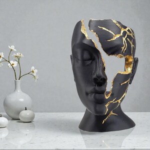 Op de afbeelding: Een zwarte sculptuur van een menselijk gezicht met een ontbrekend gedeelte, dat een goudkleurig interieur onthult. Het gezicht is glad en heeft gesloten ogen. De gouden accenten creëren een opvallend contrast. De sculptuur staat op een wit oppervlak.
