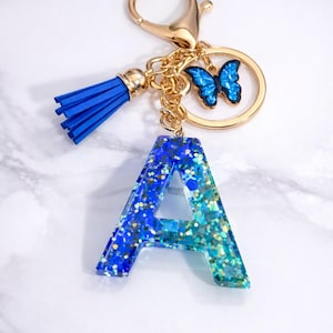 Puede incluir: Un llavero con la letra "A" de resina brillante azul y turquesa, con un cierre dorado, una borla azul y un dije de mariposa. Un accesorio personalizado.