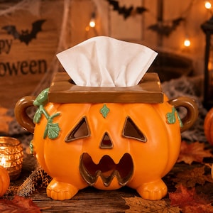 May include: A ceramic tissue box cover shaped like an orange jack-o'-lantern pumpkin with a brown lid, two side handles, and green leaf accents. The pumpkin features carved triangular eyes and a jagged mouth, with white tissues protruding from the top.
