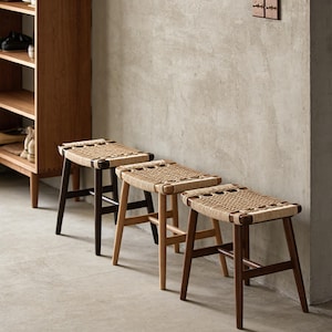 May include: Three wooden stools with woven seats. The stools are arranged in a row, with varying wood tones from dark to light brown. The seats are made of a natural-coloured woven material. The legs are angled.