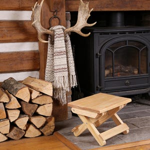 May include: A light-colored wooden folding stool sits in front of a fireplace. A stack of firewood is to the left. A scarf hangs from a set of antlers mounted on the wall. A black stove is in the background.
