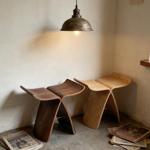 May include: Two wooden stools with a unique curved design. One stool is a dark brown color, and the other is a light tan color. The stools are in a room with a vintage-style light fixture hanging from the ceiling.