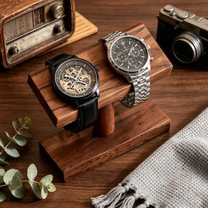 Peut inclure: Deux montres-bracelets présentées sur un présentoir en bois. L'une a un bracelet en cuir noir et un cadran squelette, l'autre a un bracelet argenté et un cadran foncé. Le présentoir est en bois foncé.