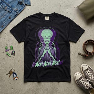 May include: A black short-sleeve t-shirt featuring a graphic of a green alien with an exposed brain, wearing a suit and holding hands together. The text "Ack! Ack! Ack!" is printed in light green and purple below the alien illustration.