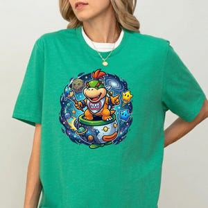 Pu&ograve; includere: T-shirt verde con una grafica a cartoni animati di un personaggio in una tazza da t&egrave;, ambientata su uno sfondo a tema spaziale. Il personaggio &egrave; arancione e giallo con un'acconciatura a cresta rossa. La tazza da t&egrave; ha un design blu e arancione.