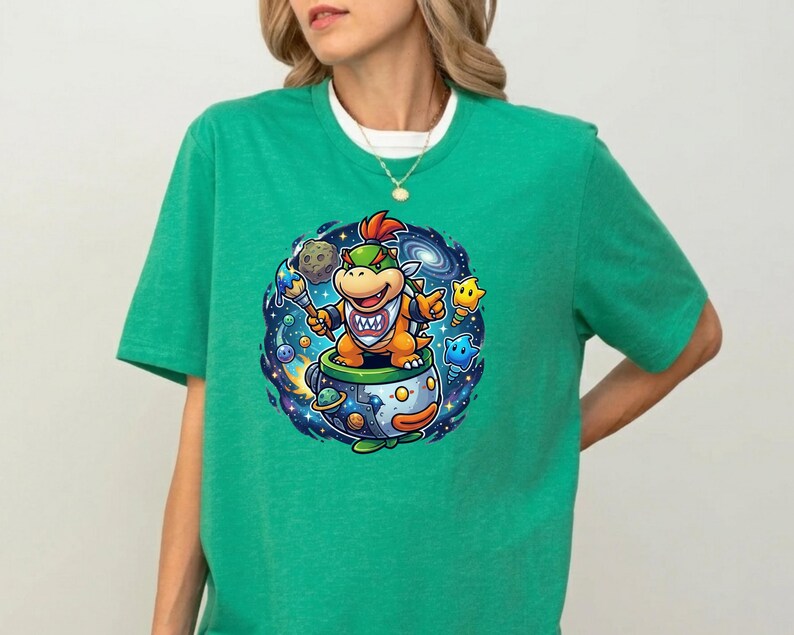 Pu&ograve; includere: T-shirt verde con una grafica a cartoni animati di un personaggio in una tazza da t&egrave;, ambientata su uno sfondo a tema spaziale. Il personaggio &egrave; arancione e giallo con un'acconciatura a cresta rossa. La tazza da t&egrave; ha un design blu e arancione.