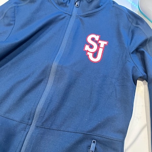 May include: Navy blue zip-up jacket with the letters "SJ" in white with a red outline on the left chest. The jacket has a high collar, a front zipper, and a zippered pocket. The jacket is made of a smooth, athletic material.