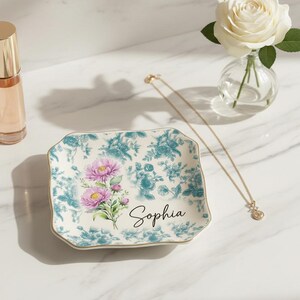 Puede incluir: Un joyero cuadrado con estampado floral azul y blanco y ribete dorado, con un ramo de flores rosas y el nombre "Sophia". También se ven un collar dorado, un frasco de perfume y una rosa blanca en un jarrón.
