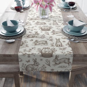 May include: A beige Easter-themed table runner with a repeating pattern of rabbits, Easter eggs, and floral elements. The runner is placed on a wooden table set with plates, bowls, wine glasses, and a vase of pink lilies.