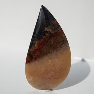 May include: A polished, teardrop-shaped gemstone. The top is black, followed by a band of red, brown, and green, leading to a tan base. The stone is displayed against a white backdrop.
