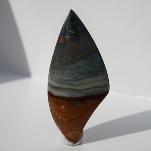 May include: A polished, teardrop-shaped stone with a layered pattern. The stone features bands of brown, grey, and blue-green hues, with a dark, glossy tip. The base of the stone is a warm, reddish-brown colour.