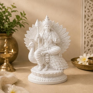 May include: White statue of a seated deity with a peacock fan, holding a spear. The statue is intricately detailed and sits on a round base. The background includes a brass oil lamp and a vase with greenery.