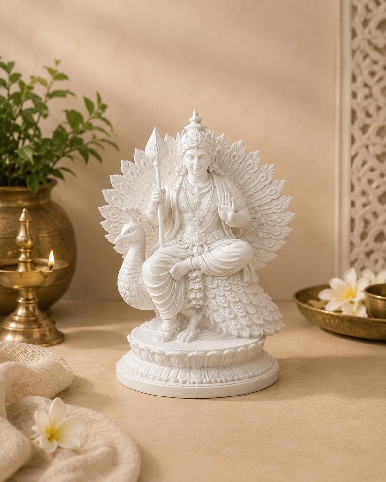 May include: White statue of a seated deity with a peacock fan, holding a spear. The statue is intricately detailed and sits on a round base. The background includes a brass oil lamp and a vase with greenery.