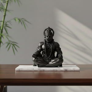 May include: A dark gray statue of a seated deity, adorned with a crown and necklace, rests on a marble slab. The statue is positioned on a dark wooden table, with bamboo plants in the background. The figure is in a meditative pose.