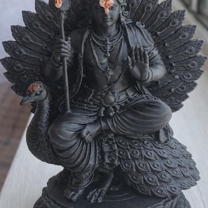May include: A black statue of a deity seated on a peacock, holding a spear. The deity wears a crown and jewelry, with a red mark on the forehead. The peacock's tail feathers are spread behind the deity.