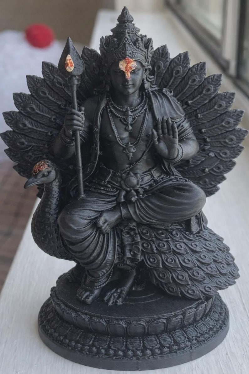 May include: A black statue of a deity seated on a peacock, holding a spear. The deity wears a crown and jewelry, with a red mark on the forehead. The peacock's tail feathers are spread behind the deity.