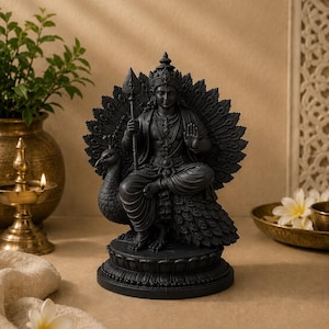 May include: A black statue of a deity seated on a peacock, holding a spear. The statue has intricate details, including a crown and jewelry. The peacock's feathers are fanned out behind the deity. The statue is on a decorative base.