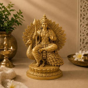 May include: A golden statue of a deity seated on a peacock, holding a spear. The statue is adorned with a crown and jewelry, set against a neutral background. The peacock's feathers fan out behind the figure.