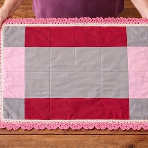 May include: A rectangular, quilted placemat with a pink crocheted border. The center features a patchwork design of red, pink, and gray squares. The placemat is held by a person's hands, showcasing the handmade quality.