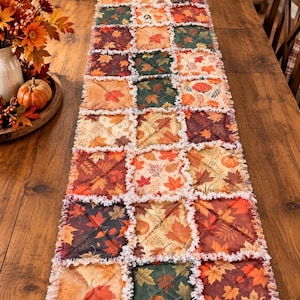 May include: A rectangular rag quilt table runner featuring a patchwork design of fabric squares with autumn-themed prints including orange, red, and dark green maple leaves and pumpkins, finished with frayed white edges and diagonal stitching.