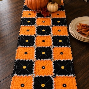 May include: A rectangular table runner featuring a chequered pattern of alternating black and orange fabric squares. Each square is quilted with an X-stitch and centred with a single button. The edges have a frayed, white-trimmed finish.