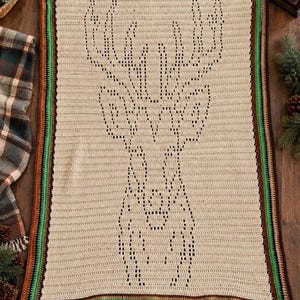 May include: A rectangular crochet blanket in a light beige color featuring a dark brown deer head silhouette created with openwork stitches. The blanket is finished with a multi-colored border in shades of brown, green, and dark brown.
