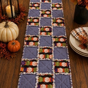 May include: A rectangular rag quilt table runner featuring a checkerboard pattern of solid periwinkle blue squares and patterned squares with orange, green, and white pumpkin motifs on a black background, finished with frayed white fabric edges.