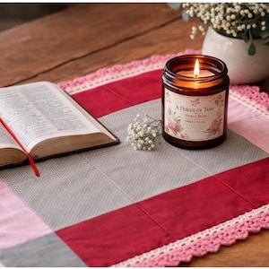 May include: A rectangular quilted mat featuring a patchwork design of grey, pink, and deep red fabric panels with a pink crocheted border. An open book with a red ribbon bookmark and a lit candle in a brown glass jar labelled A Flicker of Time sit on top.