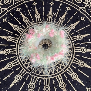 Puede incluir: Un accesorio para el cabello hecho a mano, circular, con un anillo central blanco rodeado de hilo esponjoso verde claro y rosa. El accesorio está sobre un fondo negro con un diseño celestial dorado.