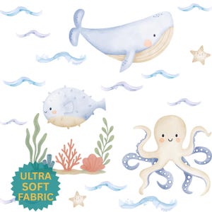May include: Watercolor illustration featuring a whale, pufferfish, and octopus. The image includes sea plants, starfish, and wavy lines. The text "ULTRA SOFT FABRIC" is on a teal tag. The color palette is soft blues, beige, and coral.