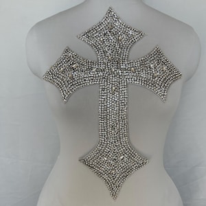 May include: A large, silver cross-shaped applique, densely covered in small, sparkling rhinestones. The cross has a detailed, ornate design with pointed edges. The applique is displayed on a white mannequin.
