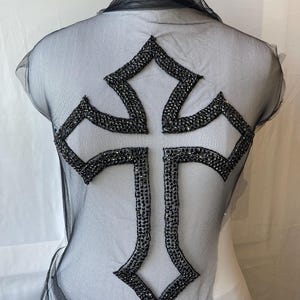 Peut inclure: Un haut en maille noire transparente présentant un grand motif de croix proéminent sur le devant. La croix est soulignée et remplie de petites perles ou strass noirs réfléchissants, créant un aspect texturé et scintillant sur le tissu.