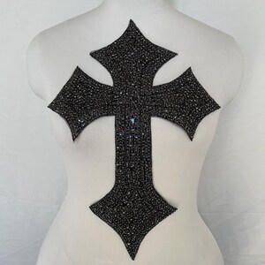 Peut inclure: Un grand écusson en forme de croix noire, densément recouvert de petits ornements scintillants. La croix est présentée sur un mannequin blanc, mettant en valeur son design et son utilisation potentielle pour les vêtements ou les accessoires. Les ornements créent un effet chatoyant.