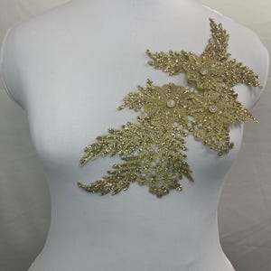 Puede incluir: Aplicación floral dorada con lentejuelas con tres grupos de flores y detalles en forma de hojas. Cada flor tiene un pedrería central. La aplicación se muestra en un maniquí blanco, mostrando su diseño y textura.