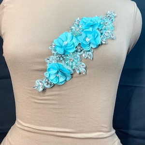 May include: A turquoise floral applique with rhinestone accents. The applique features fabric flowers and sparkling embellishments. The design is arranged in a cascading pattern, suitable for embellishing garments or accessories.