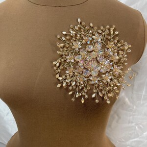 Puede incluir: Un accesorio para el cabello dorado en forma de flor con cristales transparentes e iridiscentes. La pieza está compuesta por delicadas ramas de alambre y pequeños acentos en forma de flor, creando un diseño brillante y elegante. El accesorio se muestra en un maniquí marrón.