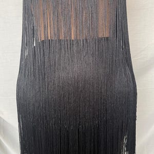 May include: A strapless black garment featuring multiple layers of long, thin, vertical fringe that covers the entire front surface. The fringe is densely packed and hangs straight down from the top edge, creating a textured, linear appearance.