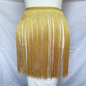 Op de afbeelding: Een goudkleurige franjerok met een brede, gestructureerde tailleband en lange, dunne, verticaal geplaatste draden in een metallic tint die een vloeiend, gelaagd uiterlijk creëren met een consistente, glinsterende textuur over het geheel.