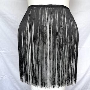 May include: A black skirt featuring a dense layer of long, thin fringe that hangs vertically from a structured waistband. The garment is composed entirely of fine, straight black strands that create a uniform, textured appearance throughout.
