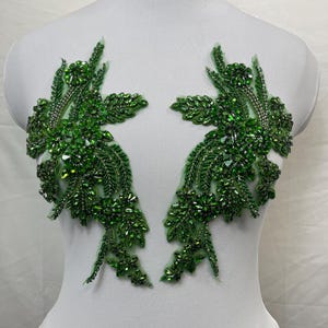 May include: A pair of symmetrical emerald green floral appliques featuring intricate beadwork, sequins, and faceted crystals. These decorative pieces are arranged on a white mannequin, showcasing detailed leaf and flower patterns in shades of green.