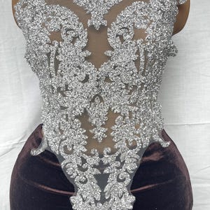 May include: Silver glitter applique with an elaborate floral and scrollwork design. The applique is set on a sheer mesh backing. The design is displayed on a brown velvet surface.