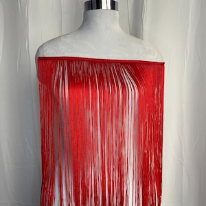May include: A vibrant red off-the-shoulder garment featuring long, flowing fringe detailing that extends from the neckline down. The piece is displayed on a mannequin, showcasing the straight, vertical alignment of the thin, silky red strands.