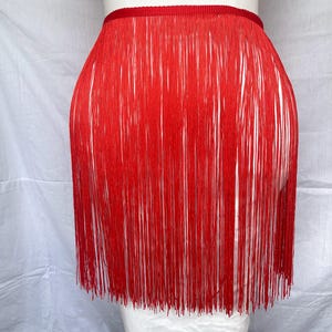 Könnte beinhalten: Ein leuchtend roter Fransenrock mit einem einfarbigen, horizontalen Bund mit gerippter Textur. Der Rock besteht aus zahlreichen langen, dünnen, vertikalen roten Strängen, die vom Bund herabhängen und ein geschichtetes, dynamisches Aussehen erzeugen.