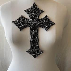Op de afbeelding: Een zwarte, met kralen bezette kruisapplicatie met een vierpuntig ontwerp. Het kruis is bevestigd aan een witte mannequin. Het kruis is gemaakt van kleine, donkere kralen en is waarschijnlijk bedoeld voor kleding- of knutselprojecten.