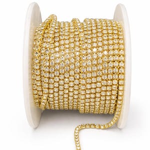 Puede incluir: Un carrete de cadena dorada con pedrería. La cadena está enrollada alrededor de un carrete de plástico blanco. Los pequeños brillantes cuadrados están muy juntos, creando una superficie continua y brillante. Ideal para la creación de joyas y proyectos de artesanía.