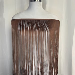 Puede incluir: Un fleco de color marrón que presenta una banda superior acanalada horizontal con largos y finos hilos verticales que cuelgan hacia abajo. El fleco se muestra sobre un torso de maniquí blanco, resaltando la textura y longitud de los hilos.