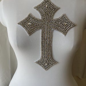 May include: A silver cross-shaped rhinestone applique. The cross is made of numerous small, sparkling rhinestones, creating a shimmering effect. The applique is displayed on a white mannequin.