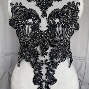 May include: A black bodice applique featuring intricate beadwork and embroidery on a sheer mesh base. The design includes dense clusters of black beads, sequins, and faceted stones arranged in a symmetrical, ornate pattern across the front.